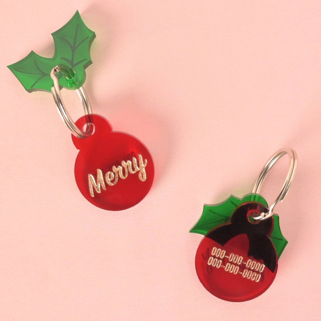 Two views (front and back) of a small, red circular pet ID tag. The tag on the left features the glitter gold slogan 'Merry,' and the tag on the right shows placeholder phone numbers. Both tags have an attached green acrylic holly leaf charm.