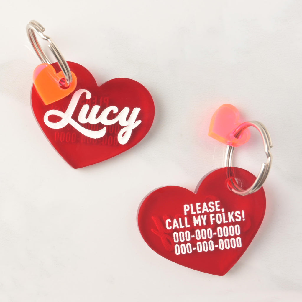 Two red heart-shaped acrylic pet ID tags with orange heart charms; one side shows the name Lucy in white script, the other side displays emergency contact information with the message 'Please, call my folks!'