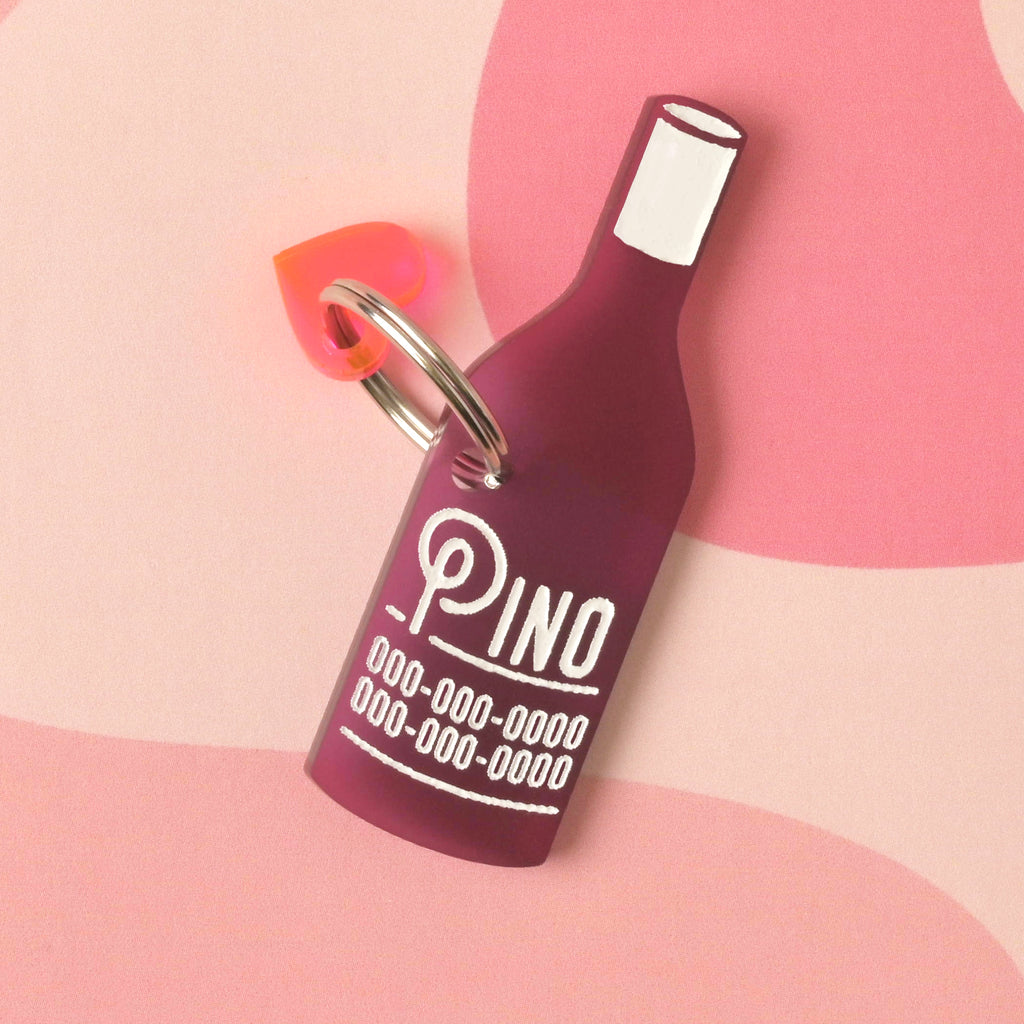 A personalized dark pink wine bottle-shaped pet tag that says "Pino" in a white groovy font, along with a placeholder phone number. The tag has a small, neon pink heart charm attached. The items are connected to a silver ring for a collar and are displayed on a pink background