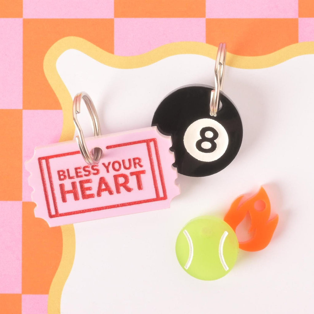 Colorful acrylic pet ID tags shaped like a “Bless Your Heart” ticket, black 8-ball, tennis ball, and flame, styled on a pink checkered background.