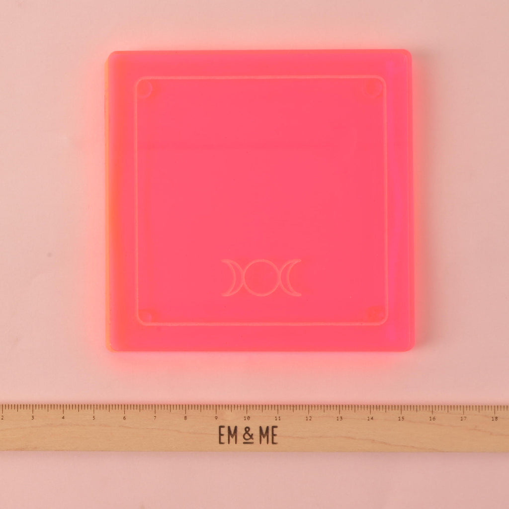 A single square, neon pink coaster is shown on a pink background. It has an etched triple moon symbol and a ruler below it with the "EM & ME" logo.