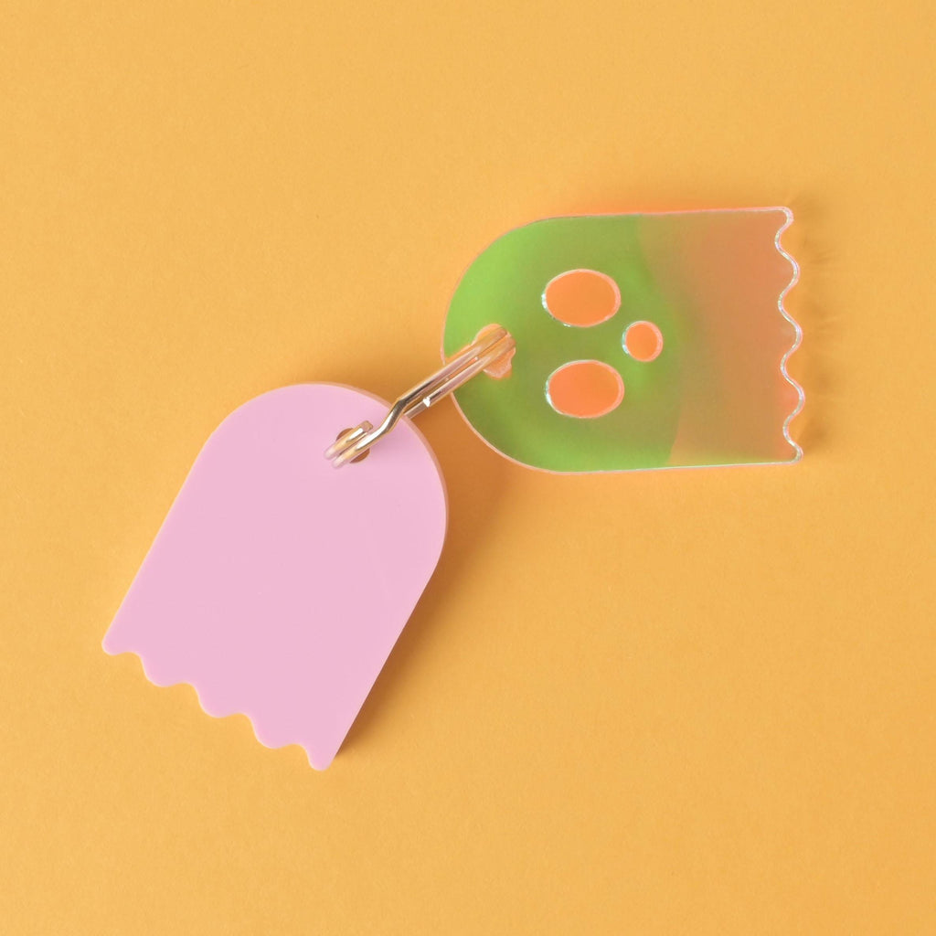 ghost-shaped pet ID tags. The tag on the left is solid light pink. The tag on the right is a translucent green with an orange glow and holographic effects. Both tags have wavy bottoms and are linked by a single silver keyring, displayed on a solid orange background.