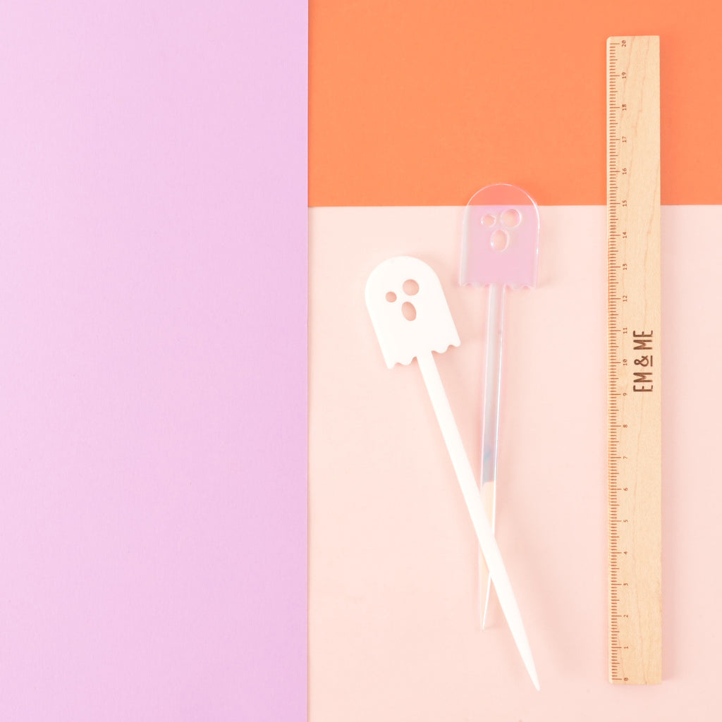 Two ghost-shaped cocktail stir sticks, one white and the other iridescent next to a wooden ruler that reads "EM & ME" on color blocked background