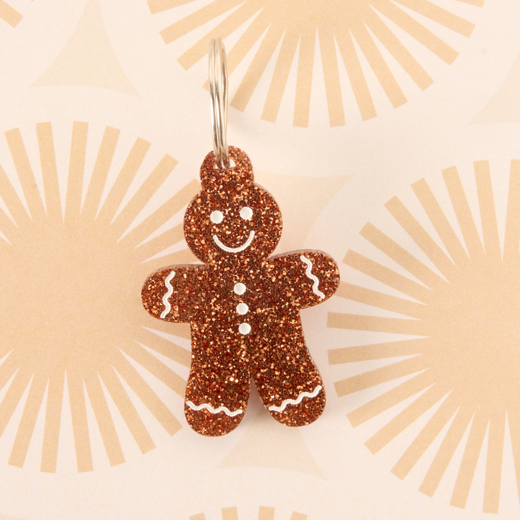 Close-up of a gingerbread man-shaped acrylic pet ID tag, featuring a brown glitter finish and white frosting details. The tag is attached to a silver split ring and displayed on a subtle patterned background with light brown sunburst/fan shapes.