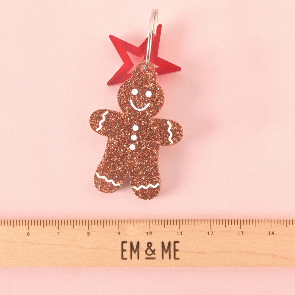 Close-up of a gingerbread man-shaped acrylic pet ID tag, featuring a brown glitter finish and white frosting details. A red star charm is attached behind the tag. The tag is displayed above a wooden ruler marked with the brand EM & ME, on a pink background.