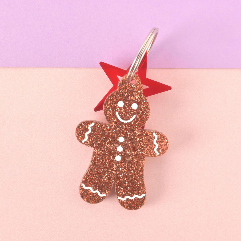 Close-up of a gingerbread man-shaped acrylic pet ID tag, featuring a brown glitter finish and white frosting details. A red star charm is attached behind the tag on the silver split ring. The tag is displayed on a pink and purple color-blocked background.