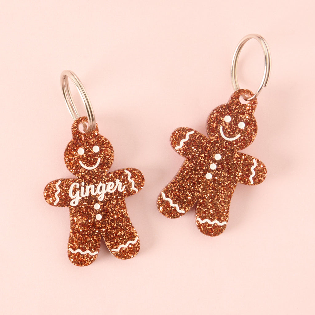 Two gingerbread man-shaped acrylic pet ID tags, featuring a brown glitter finish and white frosting details. The left tag is personalized with the name 'Ginger' on the front. The right tag shows the back view with only the frosting detail.