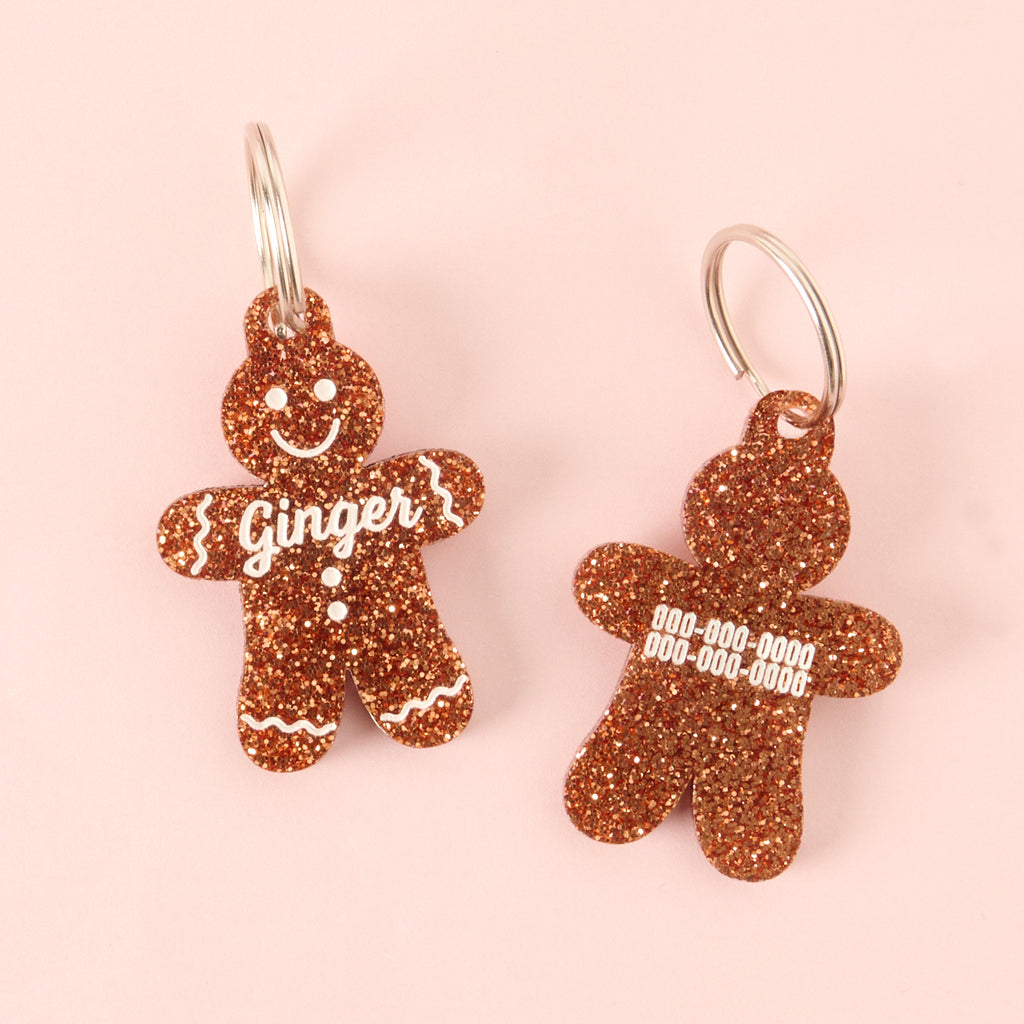 Two views of a gingerbread man-shaped acrylic pet ID tag, featuring a brown glitter finish. The left tag shows the face and the name 'Ginger' on the front. The right tag shows placeholder phone numbers on the back, with only the glitter design visible.