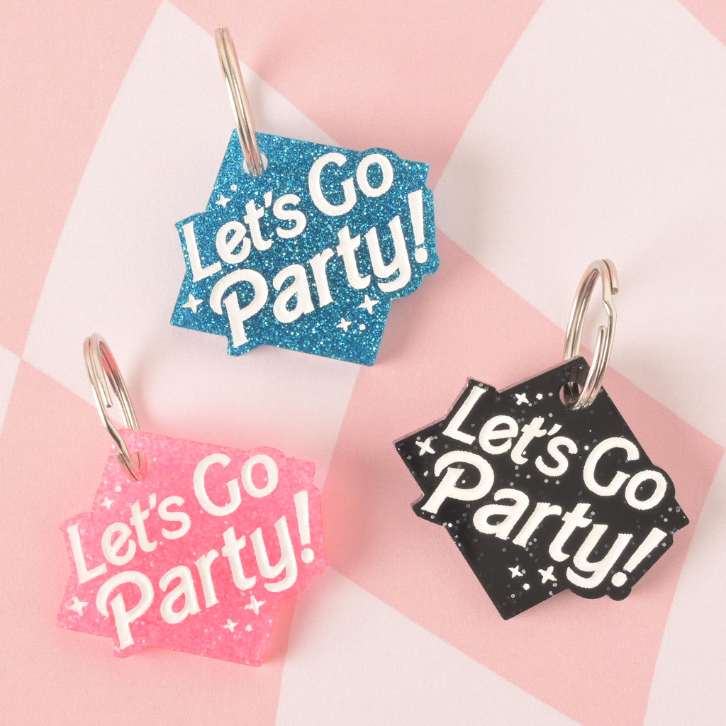 A product photo showing three glittery, rhombus-shaped pet ID tags on a checkered pink and white background. The tag on the top is blue glitter, the tag on the bottom left is pink glitter, and the tag on the bottom right is black glitter. All three tags are engraved with the words "Let's Go Party!" and have a silver split key ring.