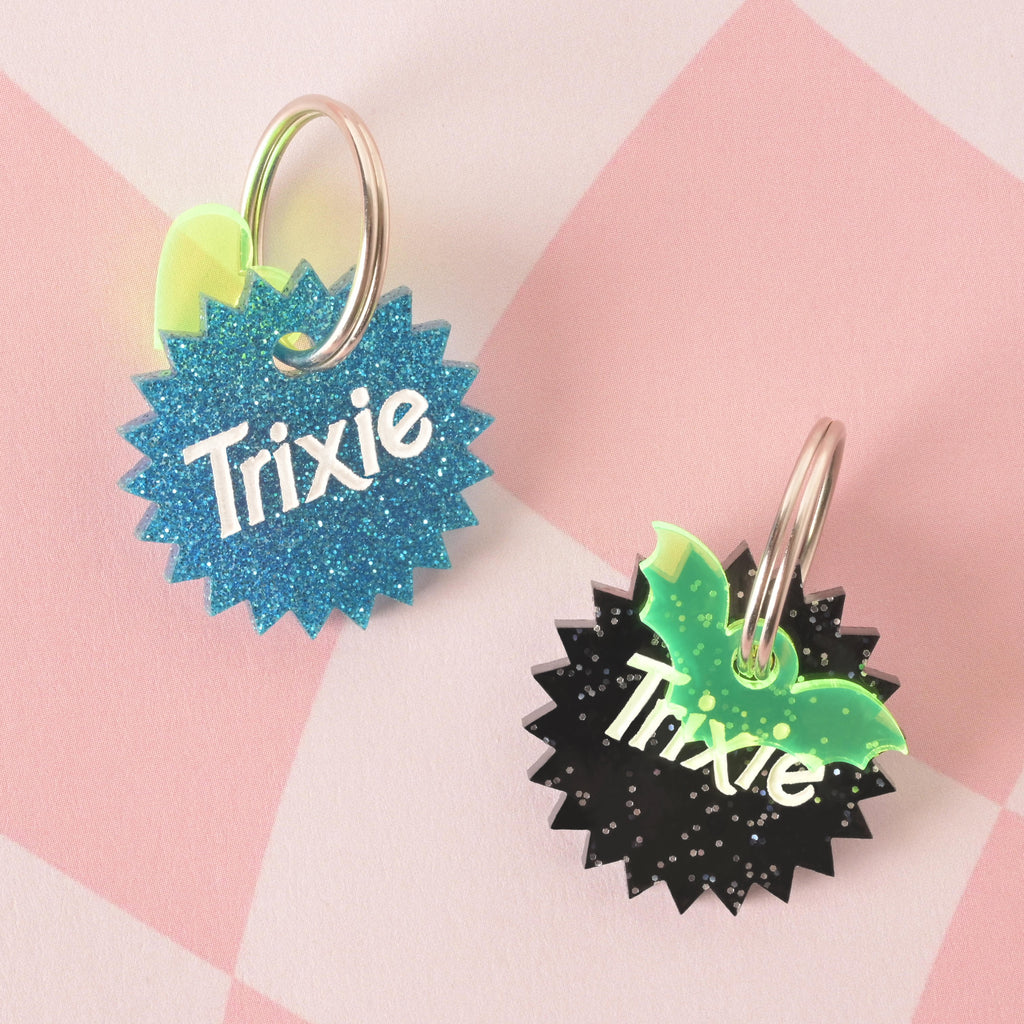 A product photo showing two unique, starburst-shaped pet ID tags on a checkered pink and white background. The tag on the left is blue glitter with a neon yellow lightning bolt charm. The tag on the right is black glitter with a neon green bat charm. Both tags are engraved with the name "Trixie" and have a silver split key ring.