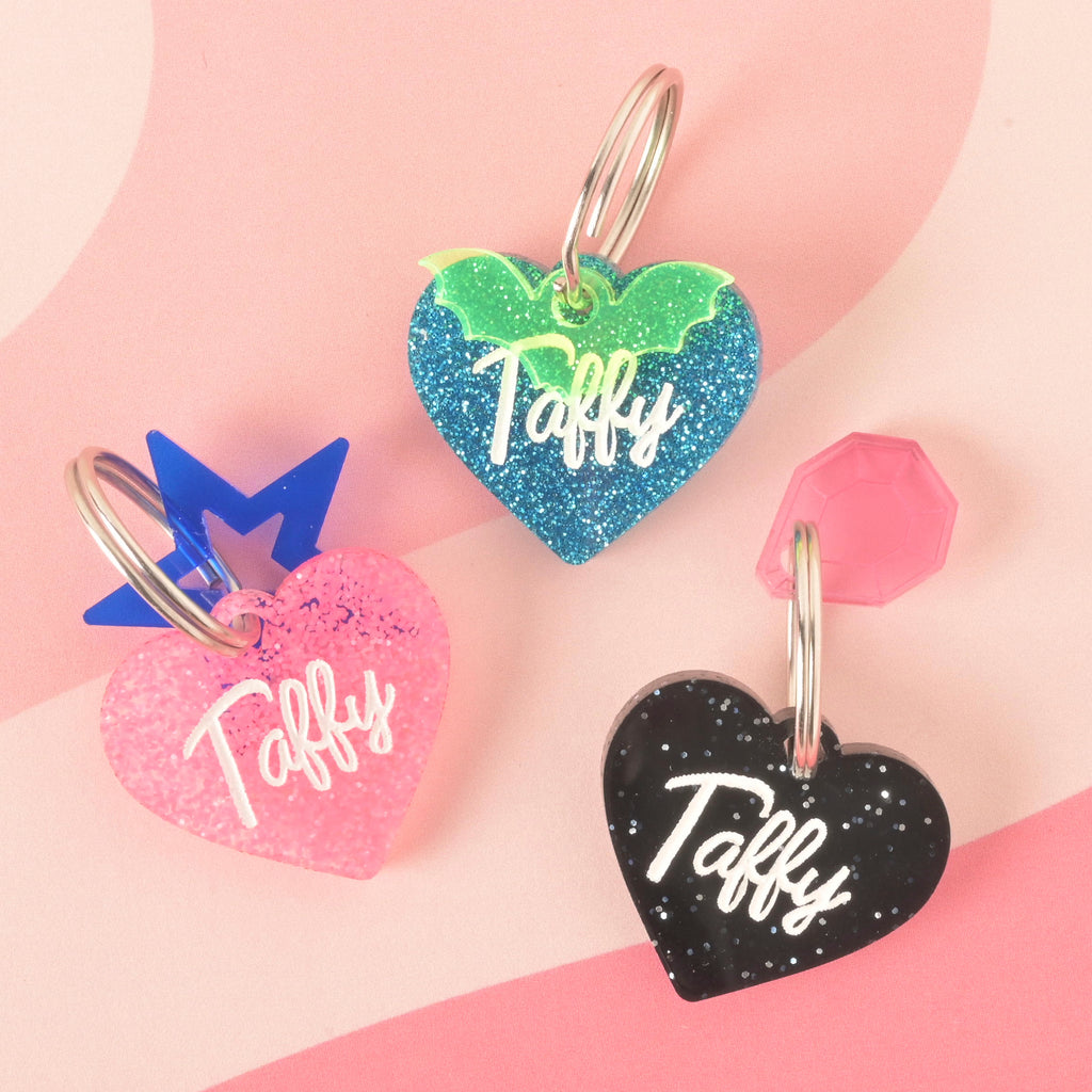 A product photo showing three heart-shaped, glittery pet ID tags on a pink background. The tag on the bottom left is pink and has a blue crown at the top. The tag on the top is blue with green glitter bat wings. The tag on the bottom right is black and has a pink gemstone charm at the top. All three tags are engraved with the name "Taffy" and have a silver split key ring.