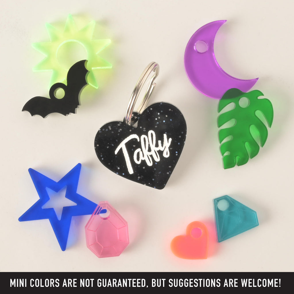 A product photo of a black glittery, heart-shaped pet ID tag with the name "Taffy" in the center. Surrounding the main tag are several small, colorful charms: a neon green sun, a black bat, a blue star, a pink faceted gem, a neon orange heart, a light blue geometric gem, a green monstera leaf, and a purple crescent moon. Text at the bottom of the image reads, "MINI COLORS ARE NOT GUARANTEED, BUT SUGGESTIONS ARE WELCOME!"