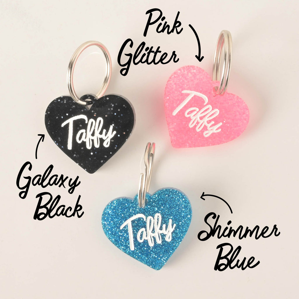 A product photo showing three heart-shaped, glittery pet ID tags. The tag on the top right is pink glitter. The tag on the top left is black glitter, labeled "Galaxy Black." The tag at the bottom is blue glitter, labeled "Shimmer Blue." All three tags are engraved with the name "Taffy" and have a silver split key ring.