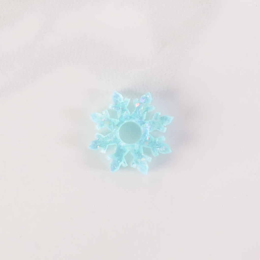 A close-up shot of a glitter teal snowflake-shaped charm with glitter accents and a hole in the middle. The charm is displayed on a white marble surface.