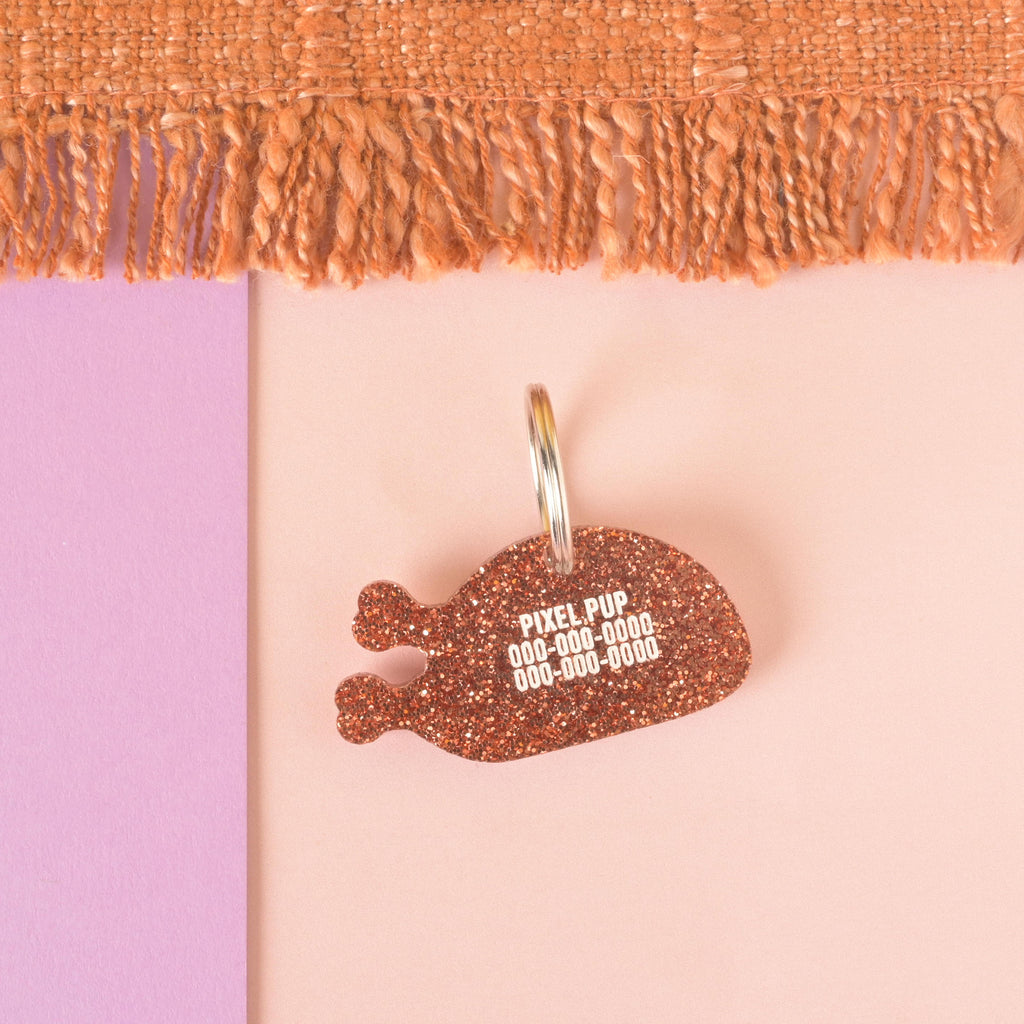 A personalized glittery copper turkey leg-shaped pet tag. The tag has the name "PIXEL PUP" and a placeholder phone number on it. The tag is attached to a silver ring, perfect for a collar, and is displayed on a pink and purple background with a brown knitted blanket at the top.