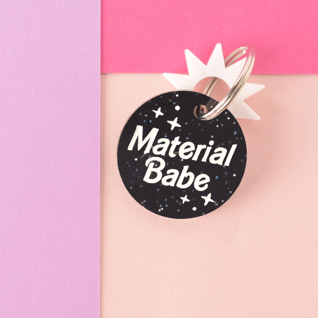 glitter black barbie inspired pet tag that reads "material babe" with a white sun-shaped mini charm on color blocked background