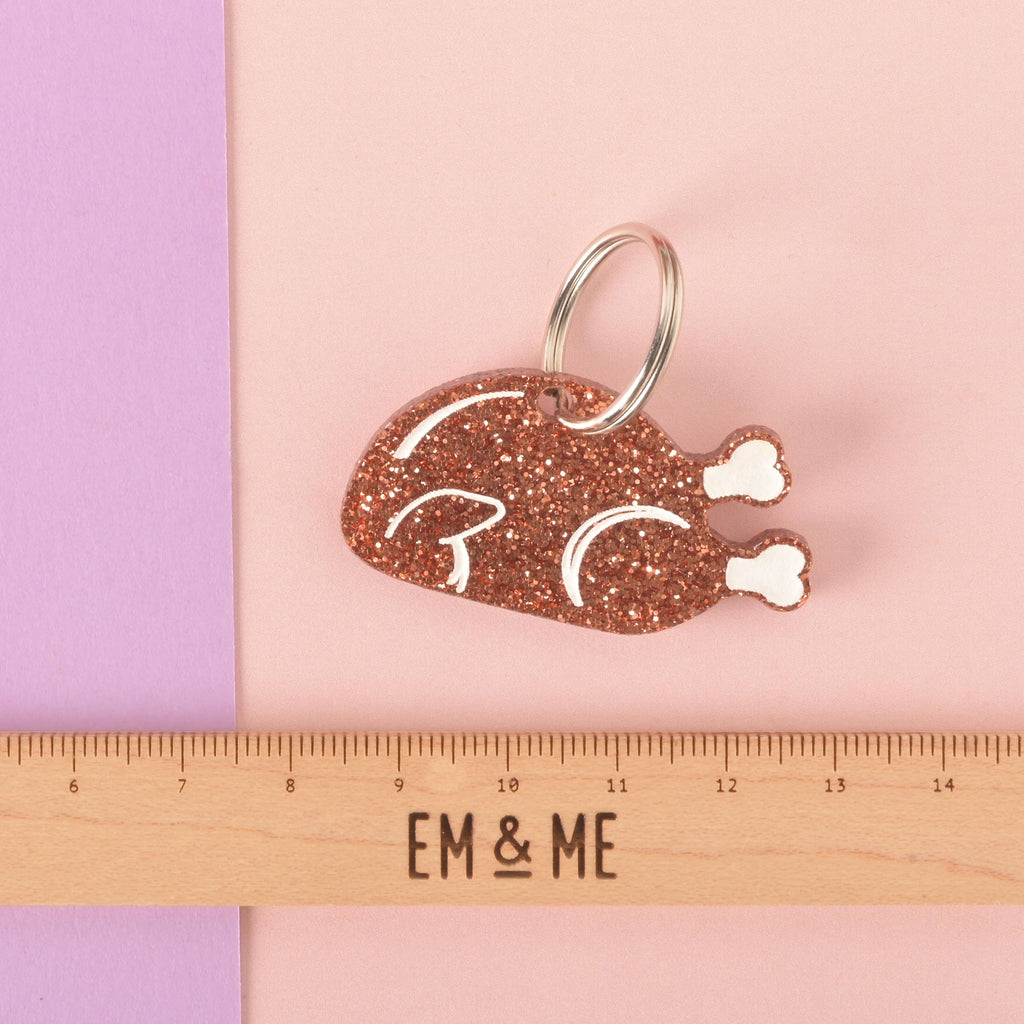 A glittery copper turkey-shaped pet tag or keychain with white bone details. The tag has a hole for attachment. A wooden ruler with the "EM & ME" logo is placed below the tag to show its size. The item is displayed on a background that is half solid purple and half solid pink.