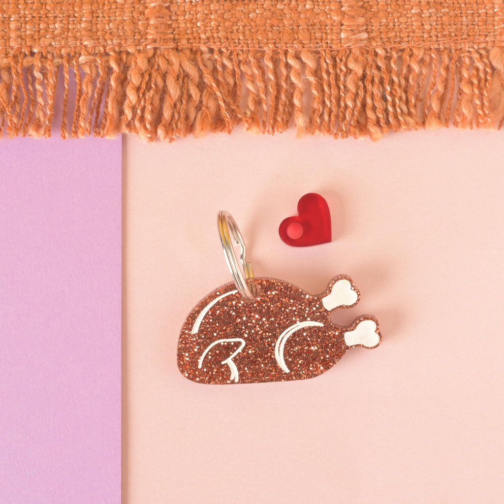 A glittery copper turkey-shaped pet tag or keychain with white bone details. The tag has a small red heart charm attached. Both are connected to a silver ring and are resting on a pink and purple background with a brown knitted blanket at the top.