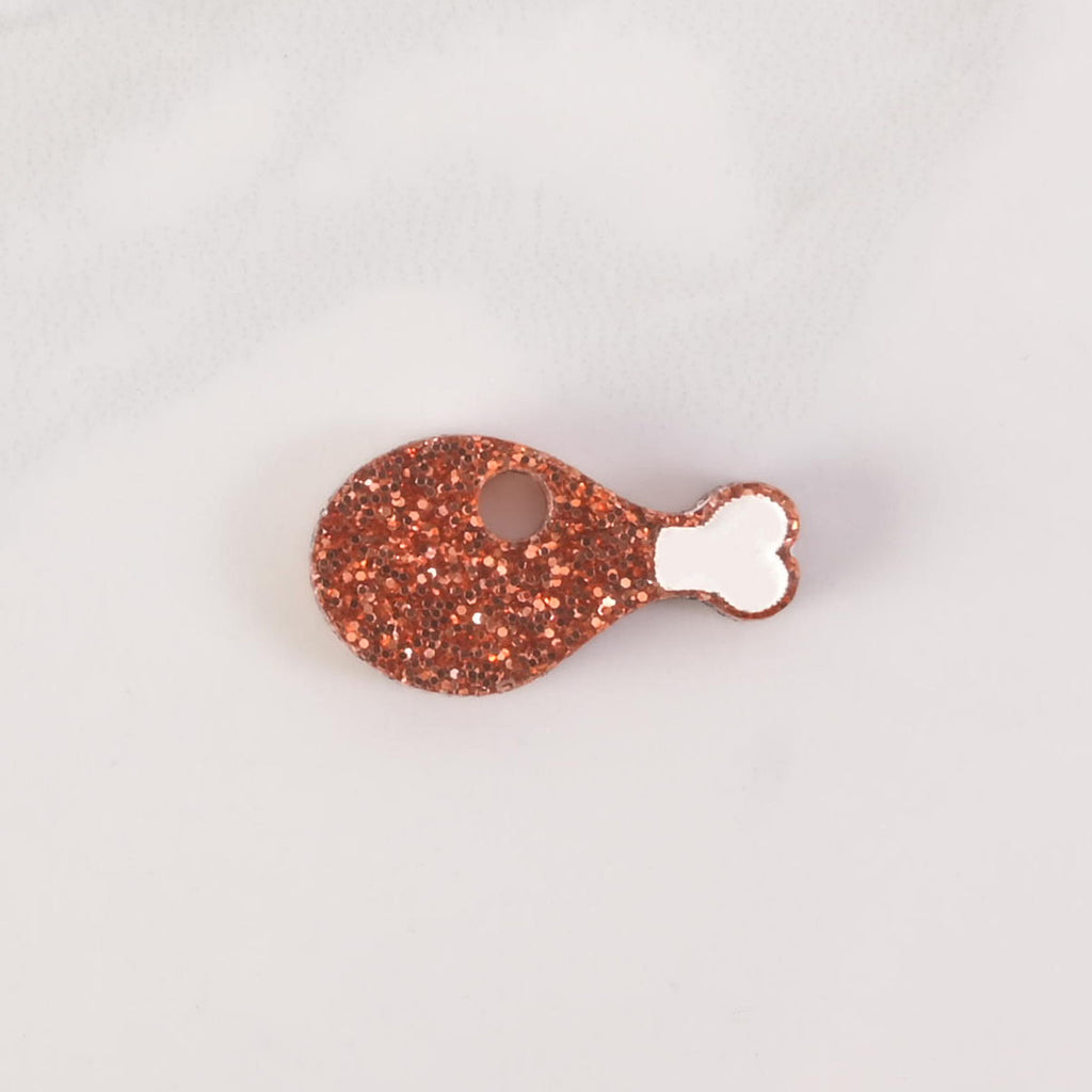 A close-up shot of a glittery copper chicken leg-shaped charm or pin. The charm has a white bone detail and a hole for attachment. The item is displayed on a white marble surface.