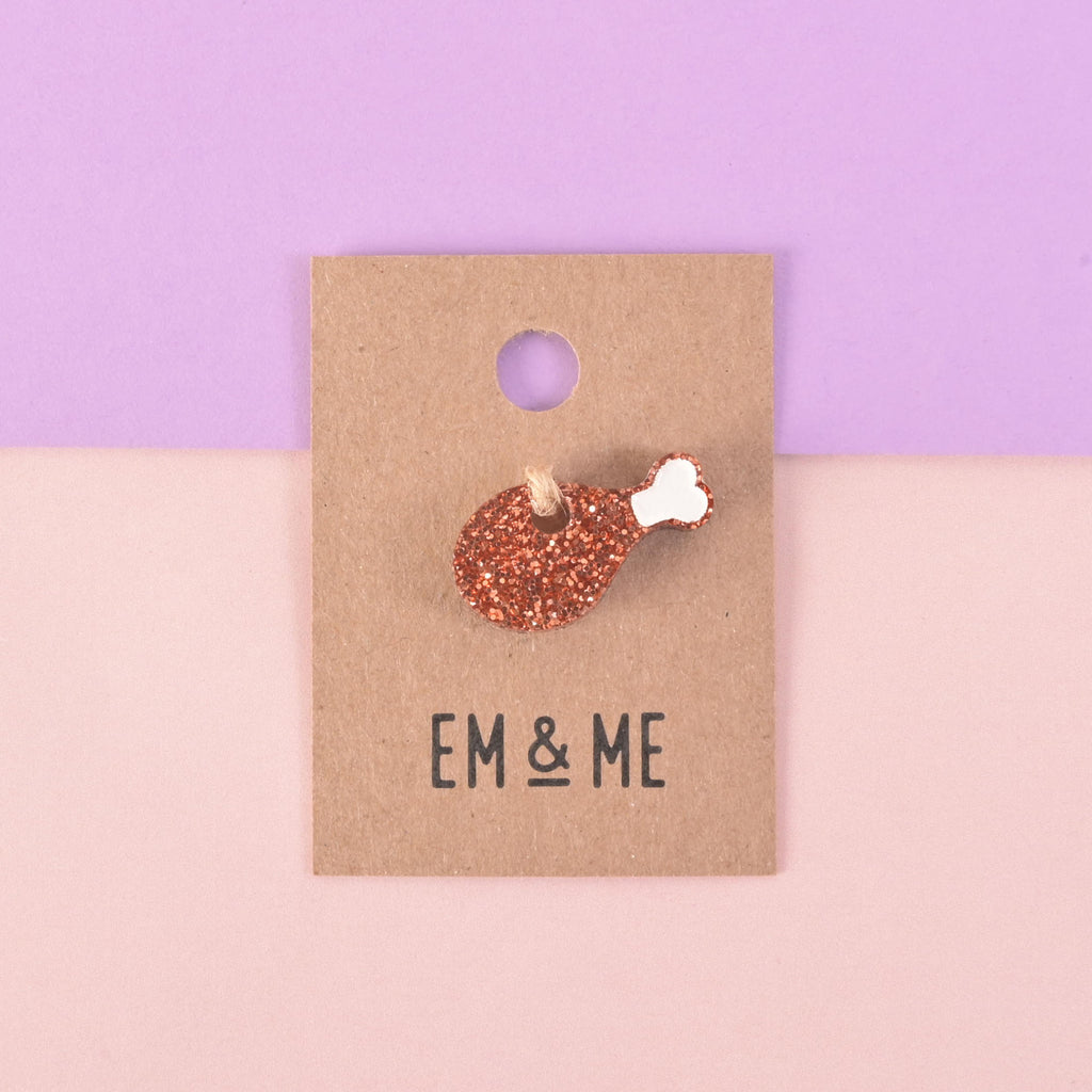 A glittery copper chicken leg-shaped charm or pin attached to a brown paper card that reads "EM & ME." The item is displayed on a background that is half solid purple and half solid pink.