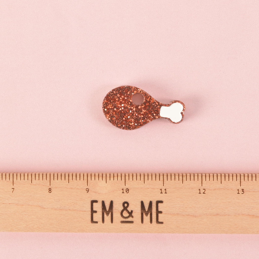 A glittery copper chicken leg-shaped charm. The charm has a white bone detail and a hole for attachment. A wooden ruler with the "EM & ME" logo is placed below the charm to show its size. The item is displayed on a solid pink background.