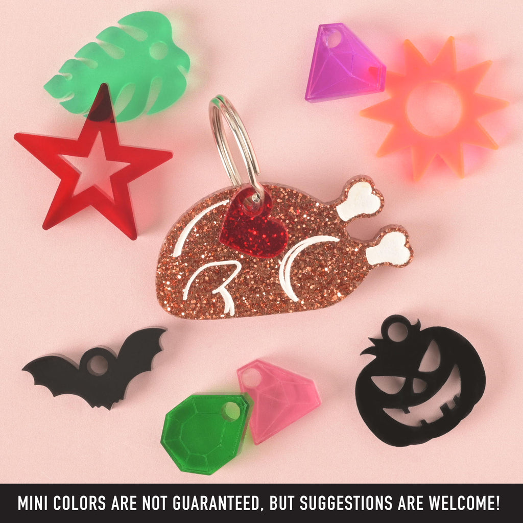 A collection of pet tags and charms scattered on a solid pink background. The central piece is a glittery copper turkey leg-shaped pet tag with a small red glitter heart charm. Surrounding it are various charms, including a red star, a green monstera leaf, a black bat, a green gemstone, a neon pink gemstone, a purple sunburst, and a black carved pumpkin. The image also contains the text "MINI COLORS ARE NOT GUARANTEED, BUT SUGGESTIONS ARE WELCOME!"