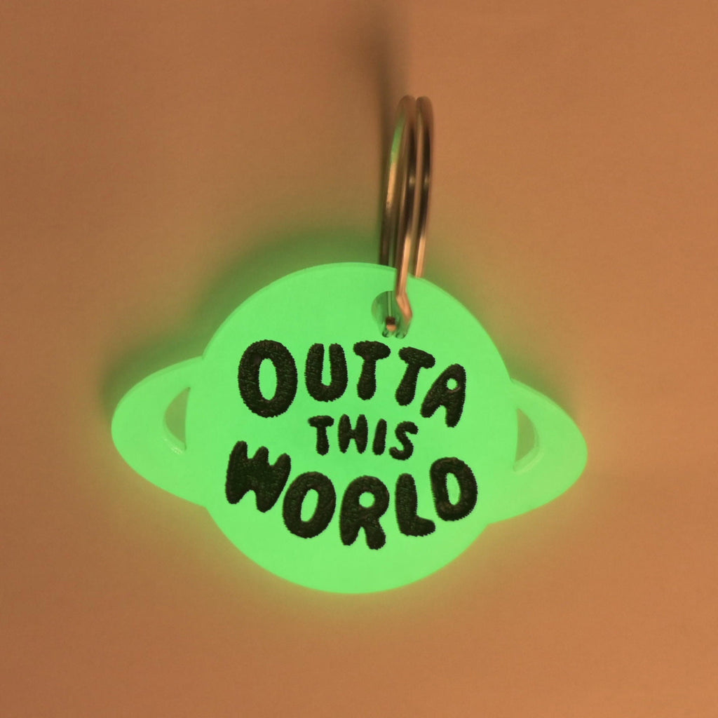 A close-up shot of a planet-shaped pet tag glowing green in a dark room. The tag is engraved with the black text "OUTTA THIS WORLD" and is attached to a shiny silver keyring.