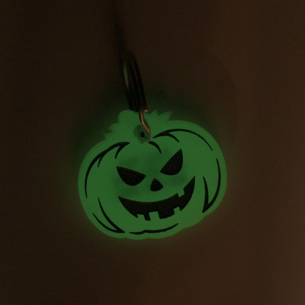 A close-up shot of a pumpkin-shaped pet tag glowing green in a dark room. The tag has a classic jack-o'-lantern face with a carved smile and triangular eyes and nose. It is attached to a silver keyring.