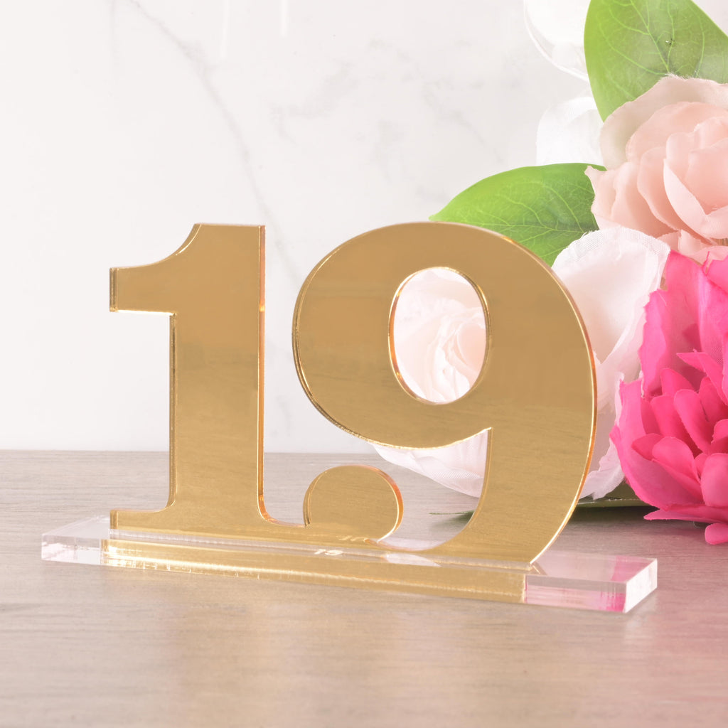 Gold acrylic table number “19” on a clear base, styled with pink and white flowers for a wedding or event centerpiece.