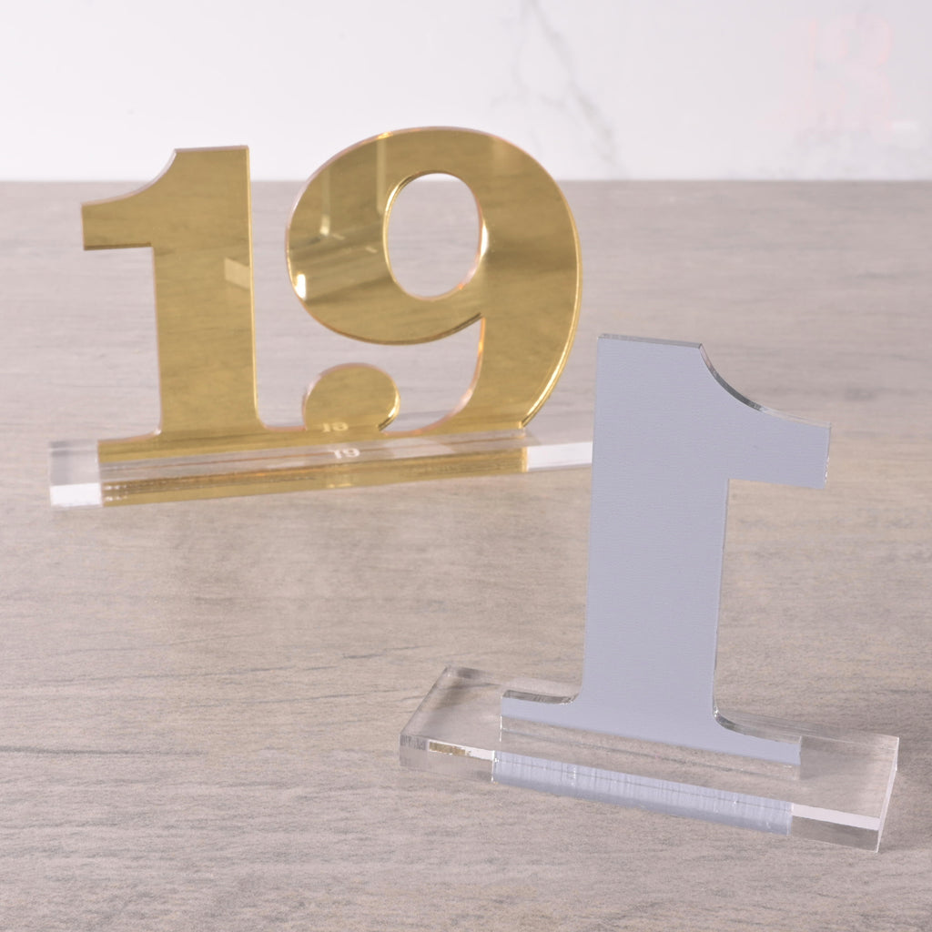 Gold acrylic table number “19” with a sleek clear base, displayed on a wooden surface for weddings or events.