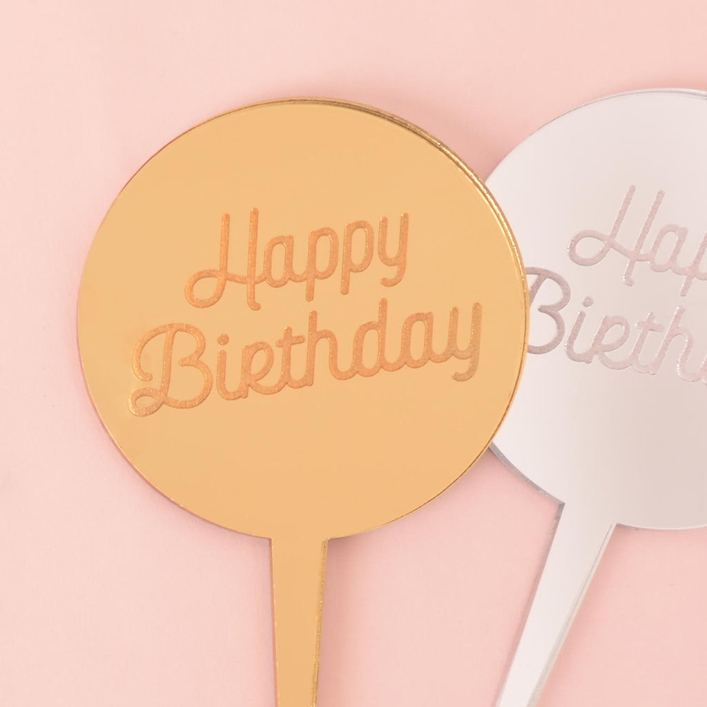 A close-up of a gold and a silver circular mirror cake topper, both with the words "Happy Birthday" etched on the surface, overlapping against a solid pink background.