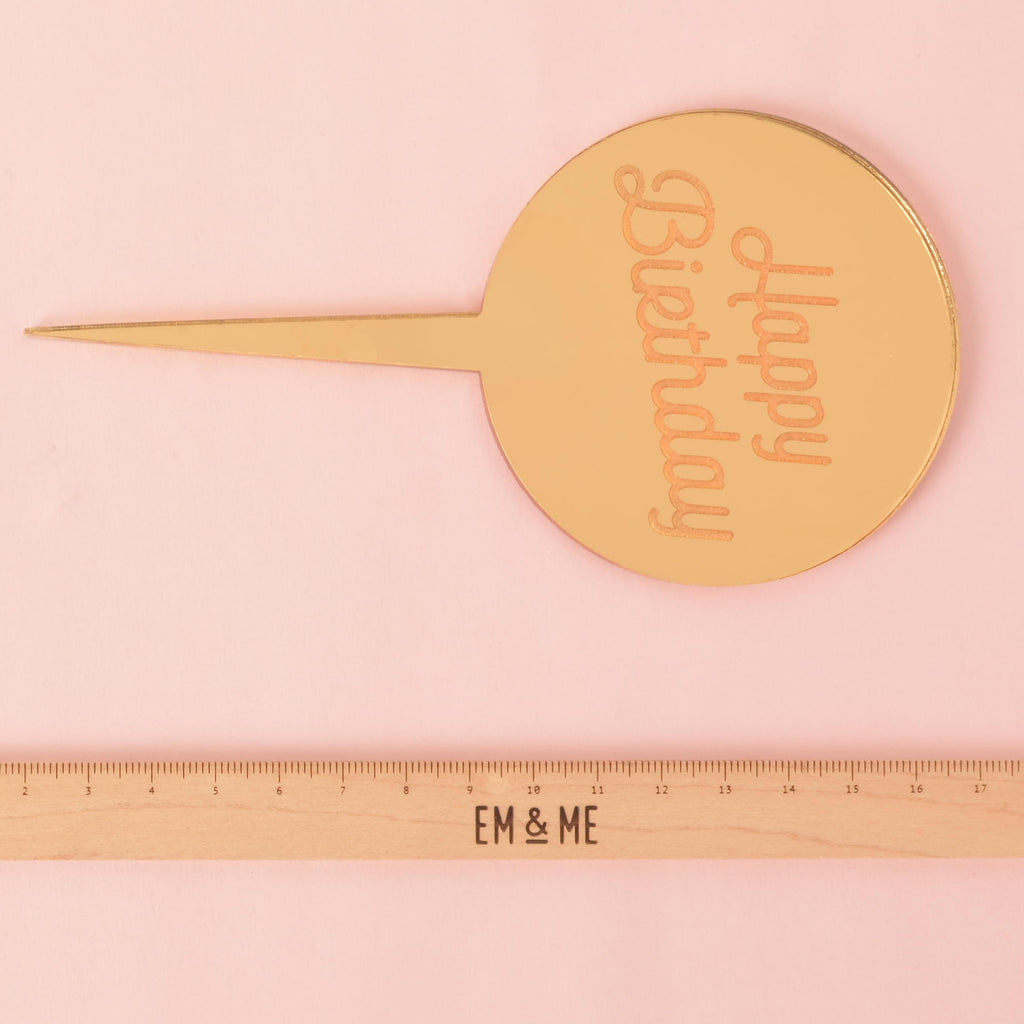 A gold circular mirror cake topper with the words "Happy Birthday" etched on the surface. A ruler with the "EM & ME" logo is placed below the topper to show its size. The background is a solid pink color.