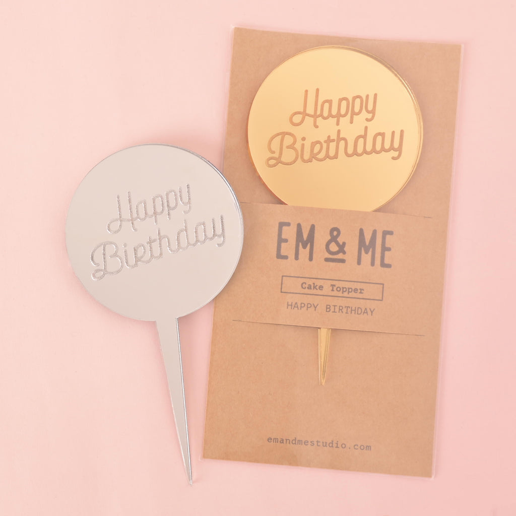A collection of "Happy Birthday" cake toppers. A silver circular mirror topper is shown on the left. A second gold circular mirror topper is sealed in its retail packaging, which has "EM & ME" printed on it. The items are displayed on a solid pink background.
