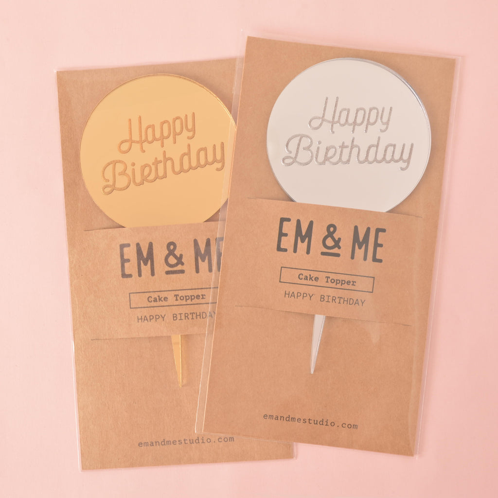 Two circular "Happy Birthday" mirror cake toppers—one gold and one silver—sealed in their retail packaging. Each brown cardstock package has "EM & ME" printed on it. The items are shown on a solid pink background.