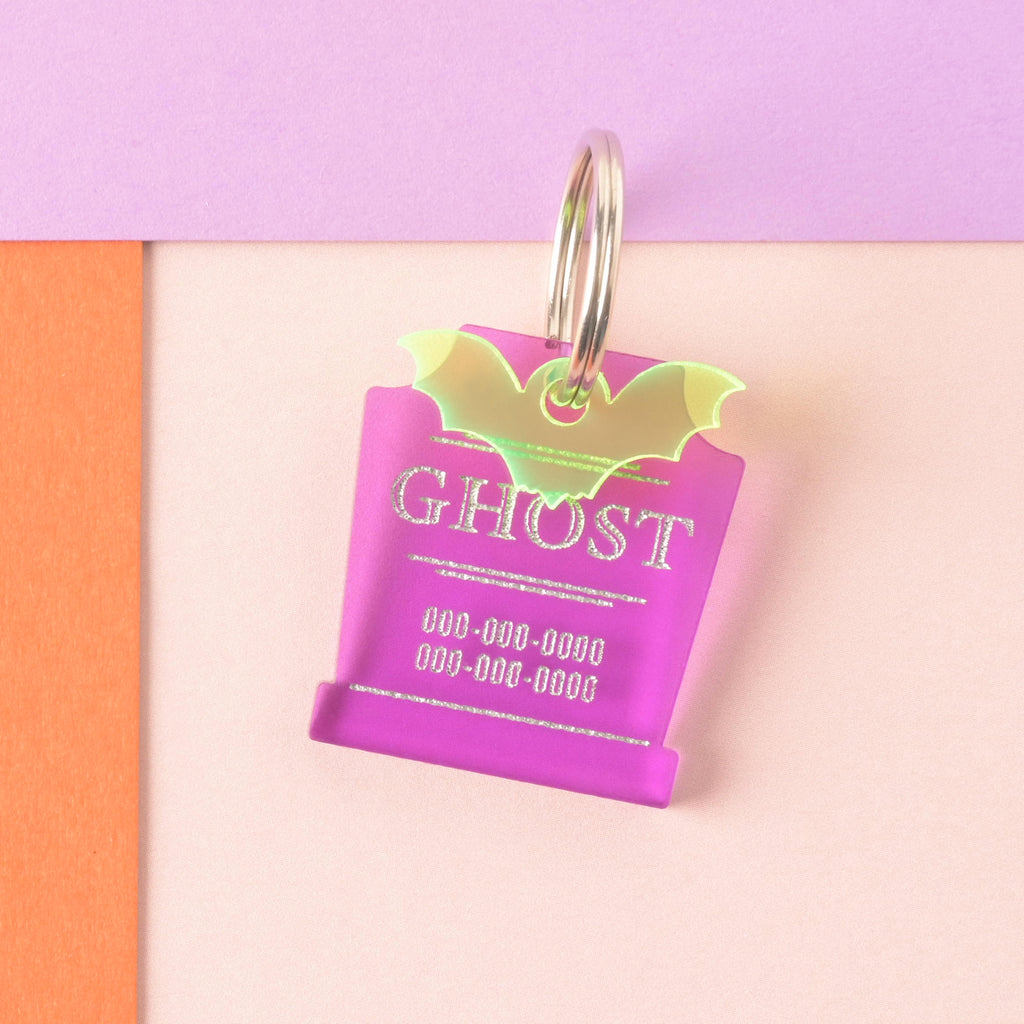 A close-up product photo of a vibrant purple tombstone-shaped pet ID tag, also known as the Grave Fabulous tag. The tag is engraved with the word "GHOST" and two lines for phone numbers. A neon yellow/green bat charm is attached to the top, which is shown with a silver split key ring.