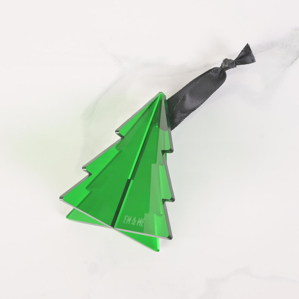 A green, flat-pack Christmas tree ornament is shown fully assembled. The ornament is made of three pieces that slide together to form a 3D shape and has a dark black ribbon tied to the top for hanging. The "EM & ME" logo is faintly etched on the front of the ornament. The item is displayed on a white marble surface.