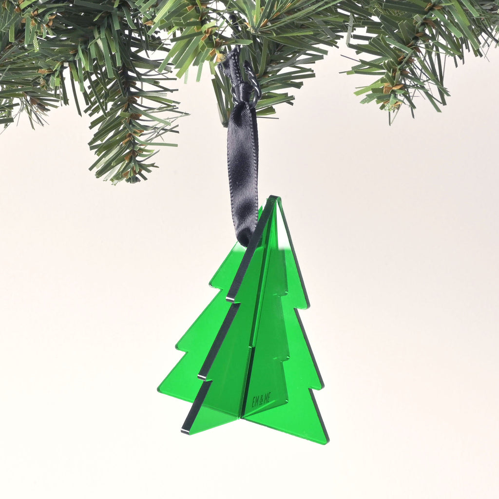 A green, flat-pack Christmas tree ornament is shown fully assembled. The ornament is made of three pieces that slide together to form a 3D shape and has a dark black ribbon tied to the top for hanging. The "EM & ME" logo is faintly etched on the front of the ornament. The item is hanging from a pine branch against a solid white background.