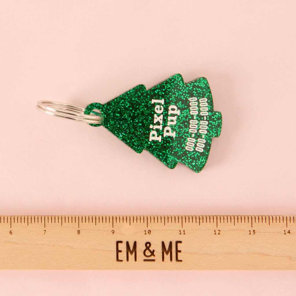 Close-up of a Christmas tree-shaped acrylic pet ID tag, featuring a dark green glitter finish. The tag is personalized with the name 'Pixel Pup' and phone numbers in white text. It is displayed above a wooden ruler marked with the brand EM & ME, on a pink background.