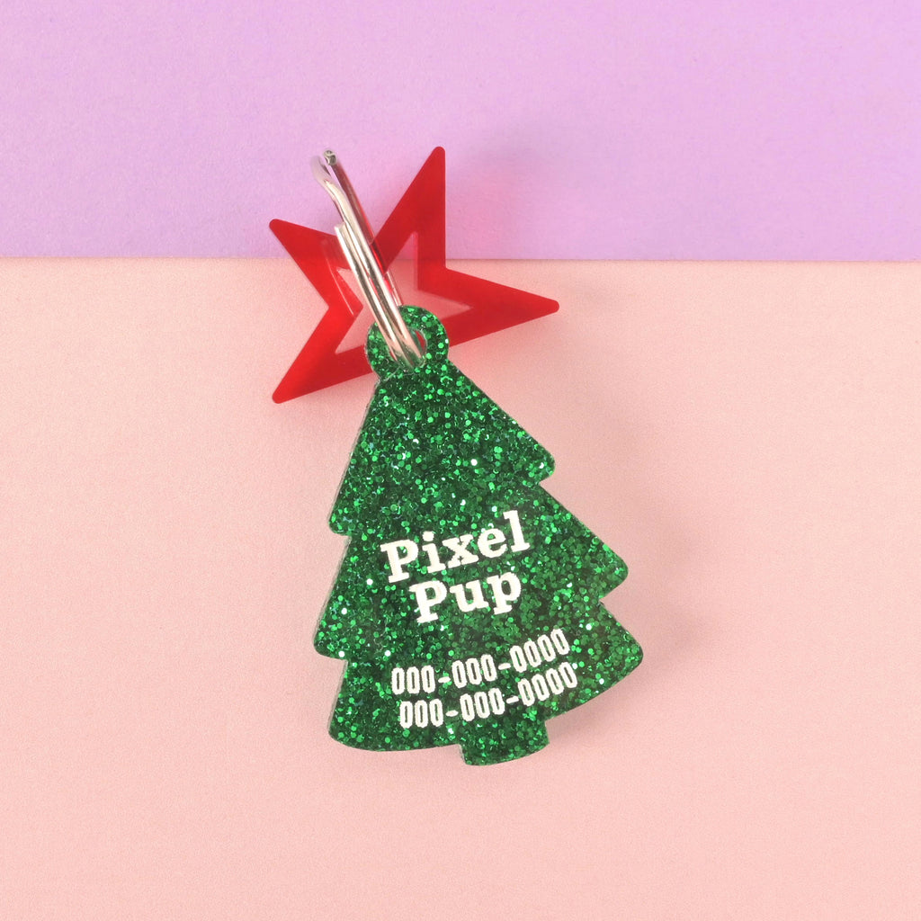 Close-up of a Christmas tree-shaped acrylic pet ID tag, featuring a dark green glitter finish. The tag is personalized with the name 'Pixel Pup' and phone numbers in white text. A translucent red star charm is attached behind the tag. The tag is displayed on a color-blocked background of light pink and purple.
