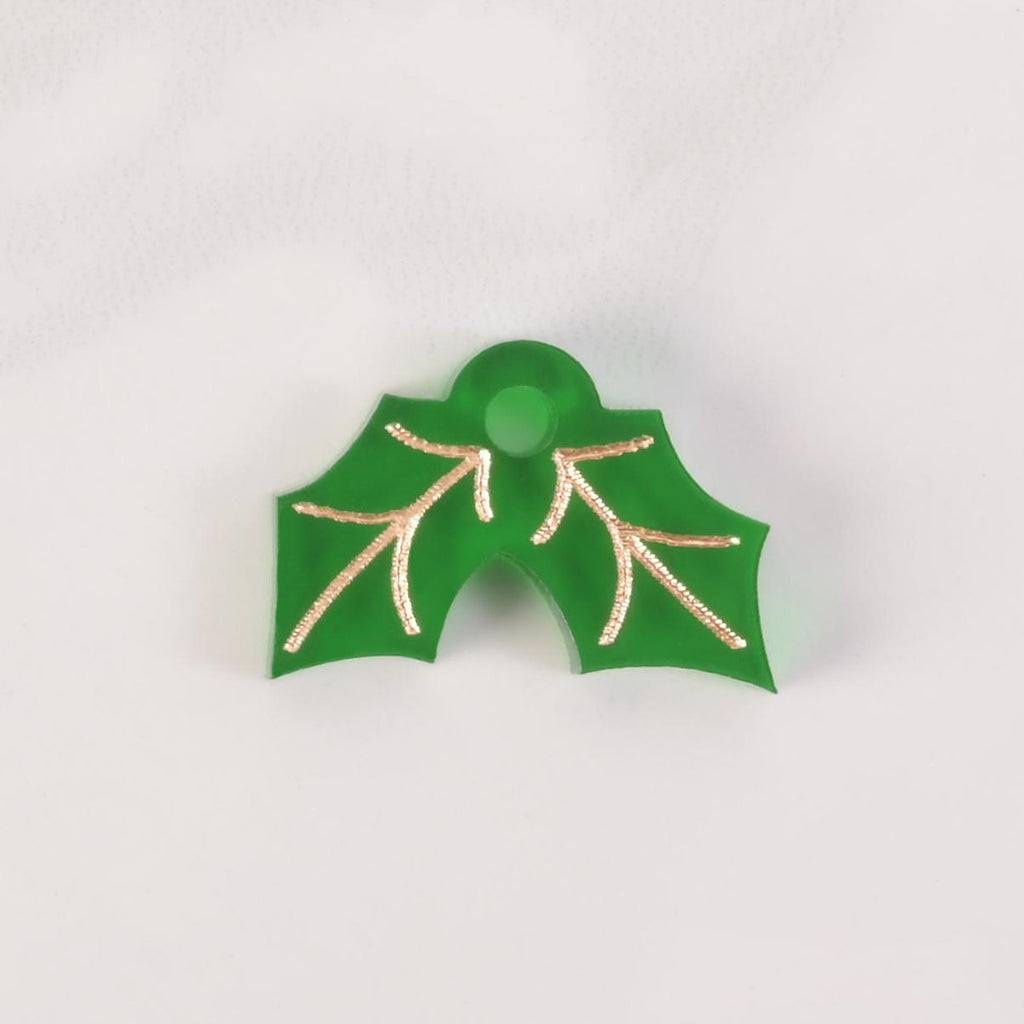 A green holly leaf-shaped charm with gold details and a hole for attachment. The item is shown on a white marble surface.