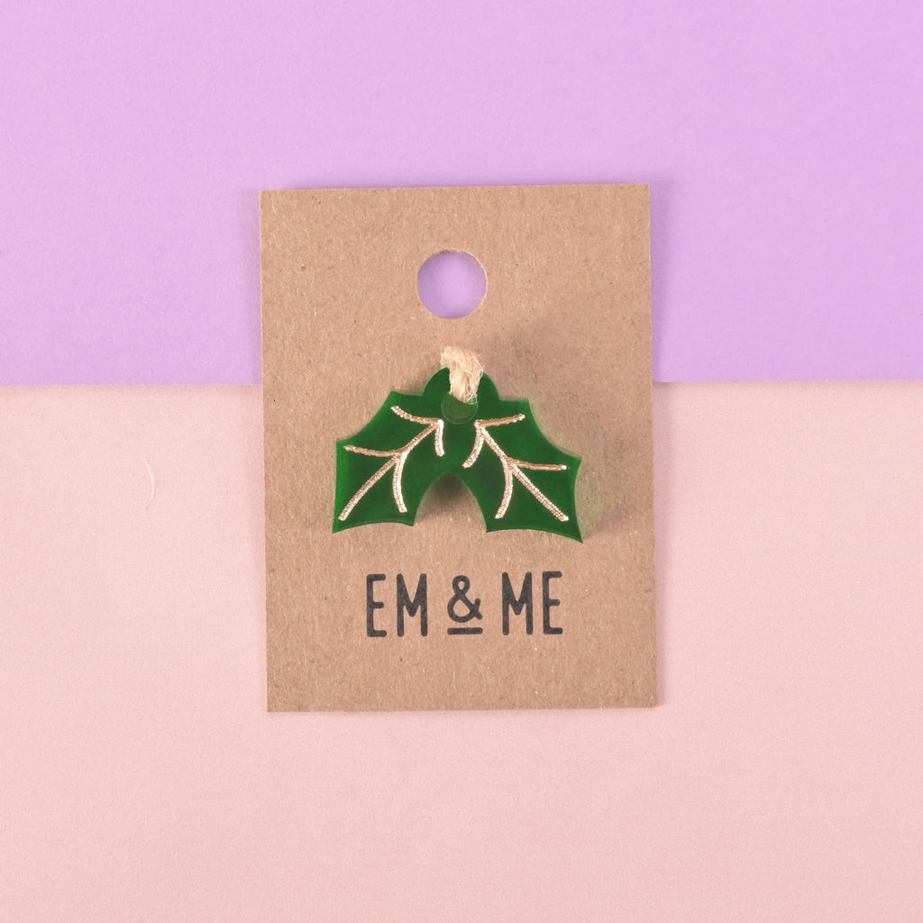 A green holly leaf-shaped charm or pin with gold details attached to a brown paper card that reads "EM & ME." The item is displayed on a background that is half solid purple and half solid pink.
