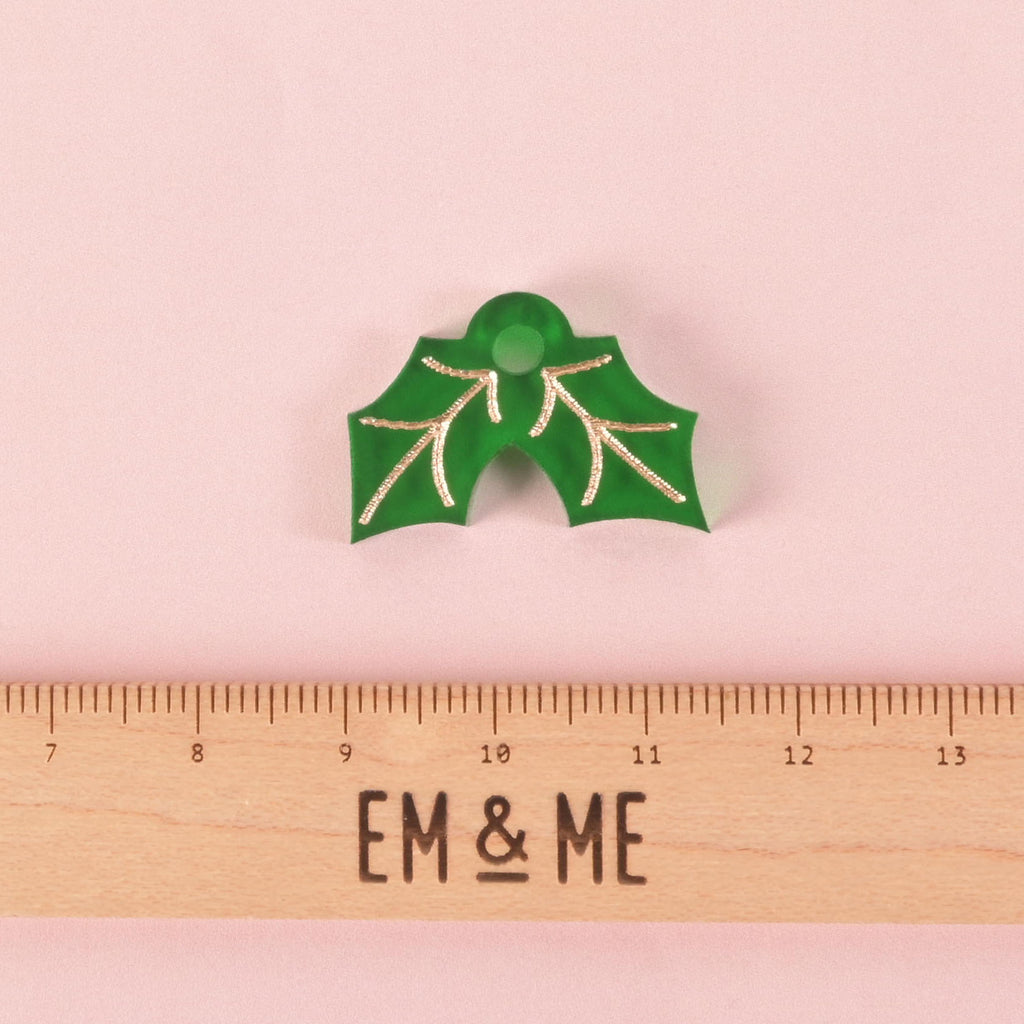 A green holly leaf-shaped charm with gold details and a hole for attachment. A wooden ruler with the "EM & ME" logo is placed below the charm to show its size. The item is displayed on a solid pink background.

