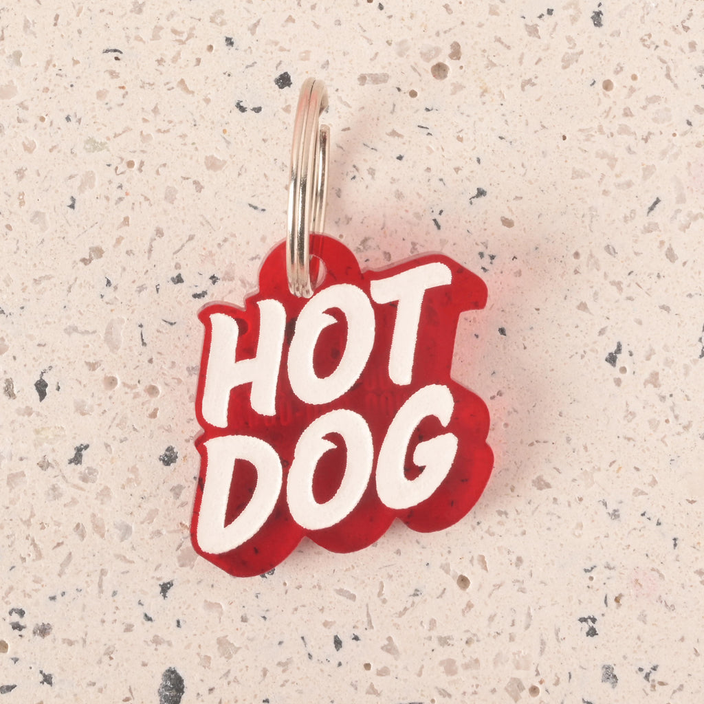 A close-up of a red pet tag in the shape of a speech bubble that says "HOT DOG" in a white groovy font. The tag is attached to a silver ring and is resting on a light-colored, speckled surface.