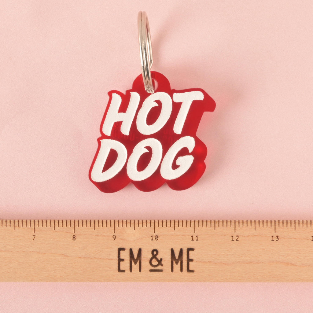 A red pet tag in the shape of a speech bubble that says "HOT DOG" in a groovy font. A ruler with the "EM & ME" logo is placed below the tag to show its size. The tag is shown on a solid pink background.