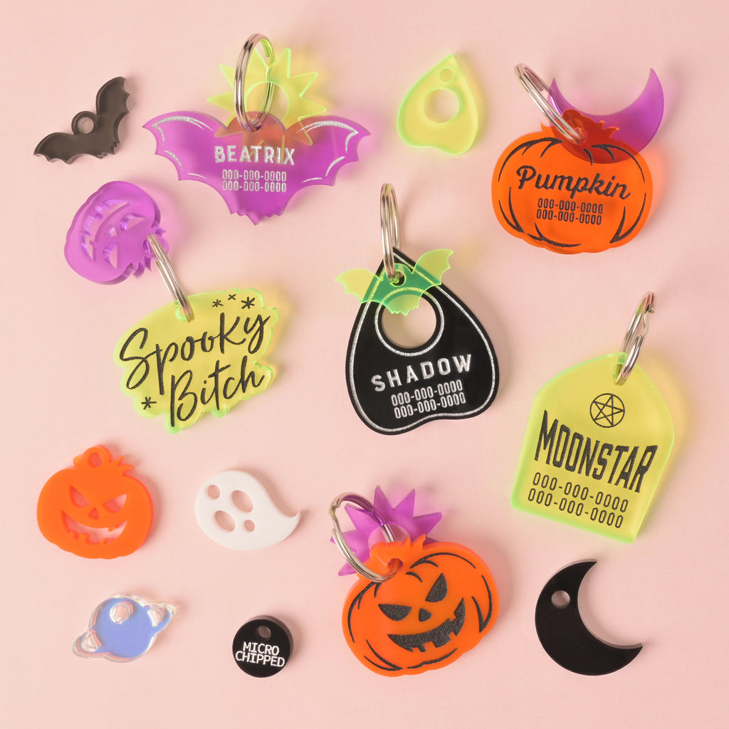 Collection of Halloween themed personalized pet tags. Purple bat, orange pumpkin, fluorescent green tombstone, orange jack-o-lantern, black ouija planchet and fluorescent green tag that reads "spooky Bitch" surrounded by various mini charms