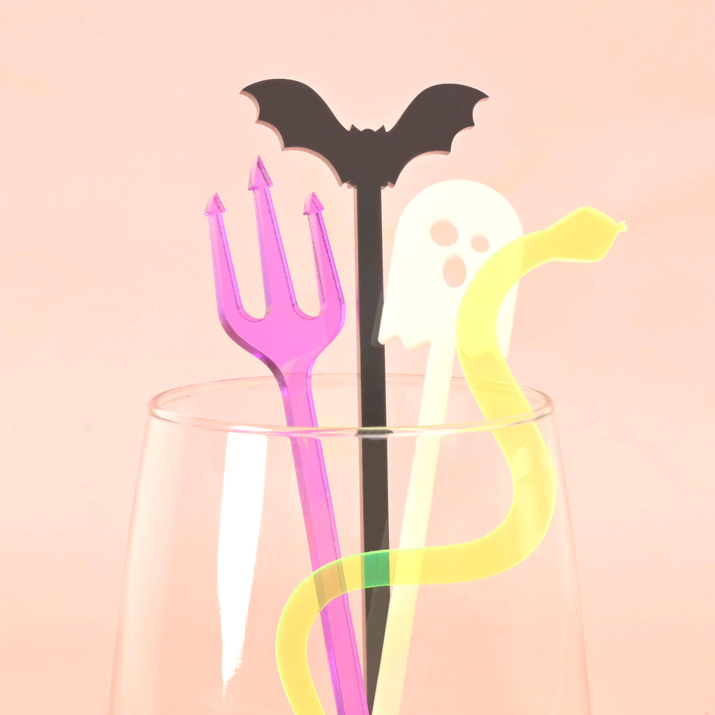 close up of 4 halloween themed stir sticks in a glass. Purple pitchfork, black bat, white ghost and fluorescent green snake on solid pink background.