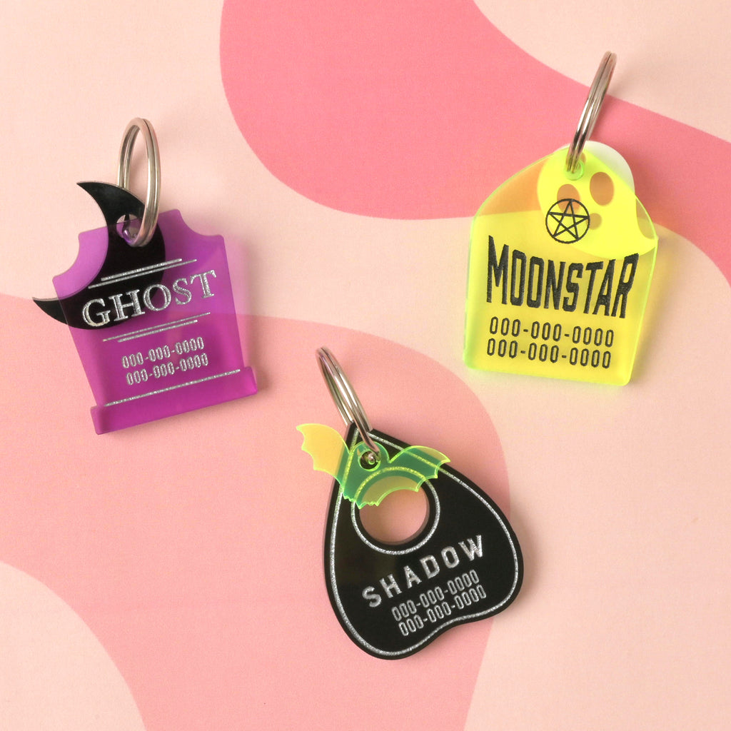 Collection of three halloween themed pet Tags, Purple gravestone-shaped pet tag with a black moon mini charm, fluorescent green tombstone-shaped pet tag with a while ghost mini charm and a black planchet-shaped pet ID tag with a fluorescent green bat mini on a wavy pink background