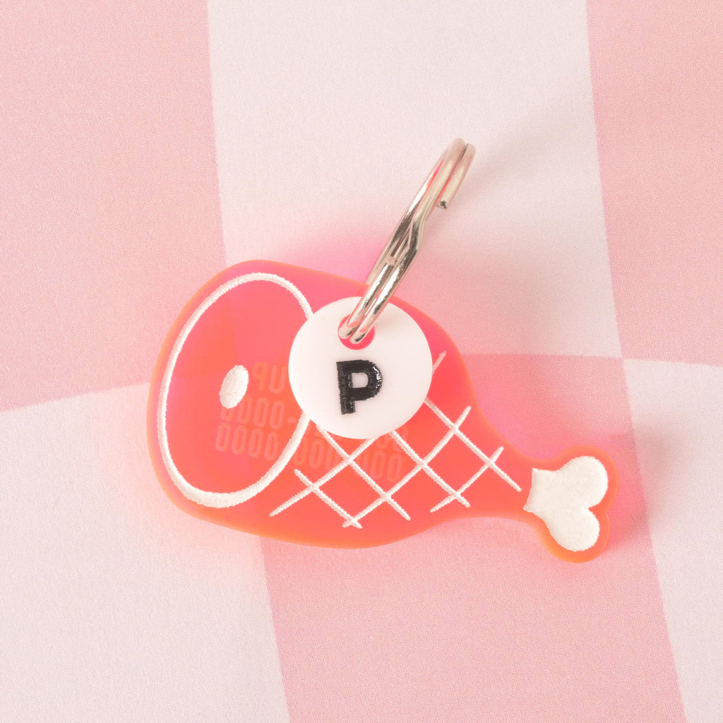 A custom, pink, ham-shaped pet ID tag with a white engraved grid pattern and bone. The tag is also engraved with a phone number that is hard to read. A small, white, circular charm with a black letter "P" is attached to the shiny silver keyring. Both charms are displayed on a pink and white checkered background.
