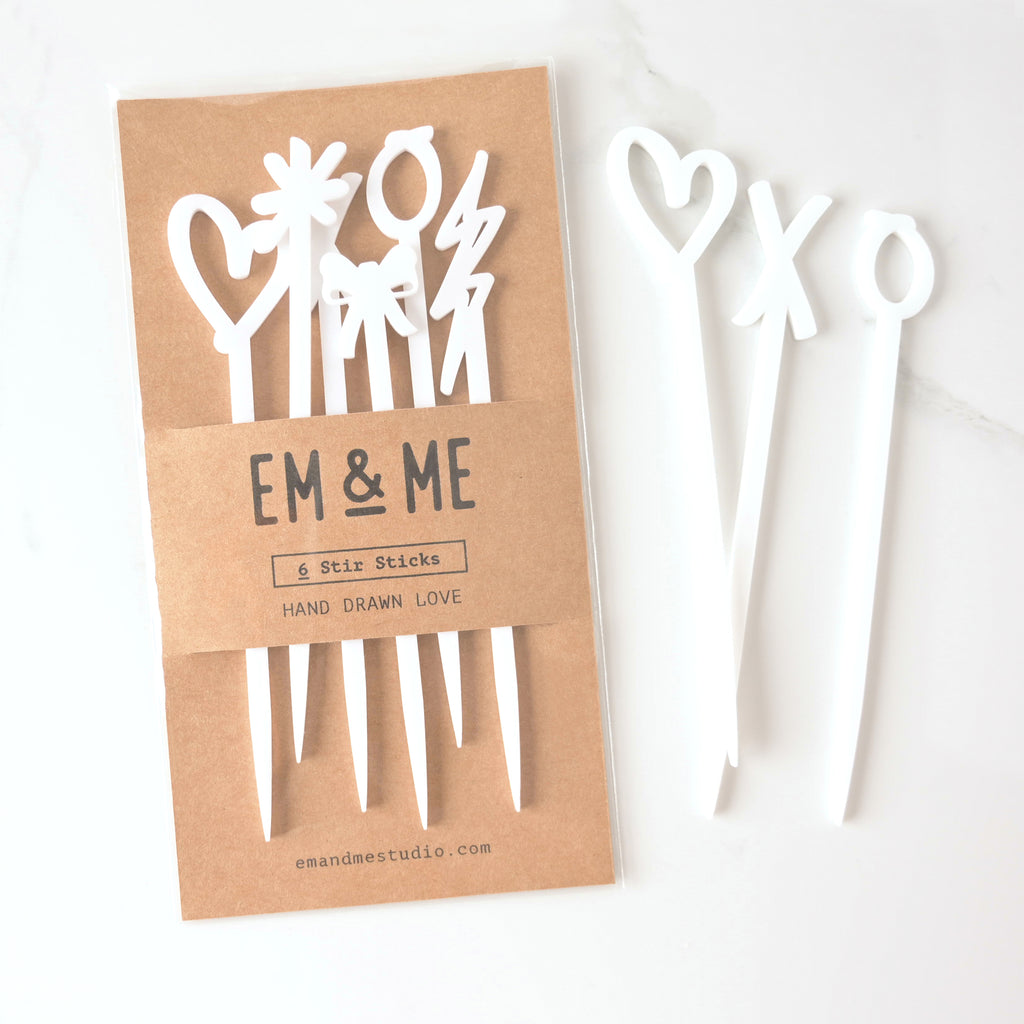 Set of 6 hand drawn love cocktail stirrers packaged in brown kraft paper with three stir sticks off to the side. Heart, X and O shapes on Marble background.