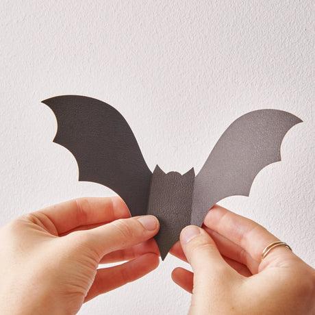 A close-up shot of two hands holding a black paper bat decoration against a white wall. The bat has its wings folded slightly to create a 3D effect. A gold ring is visible on one of the fingers.